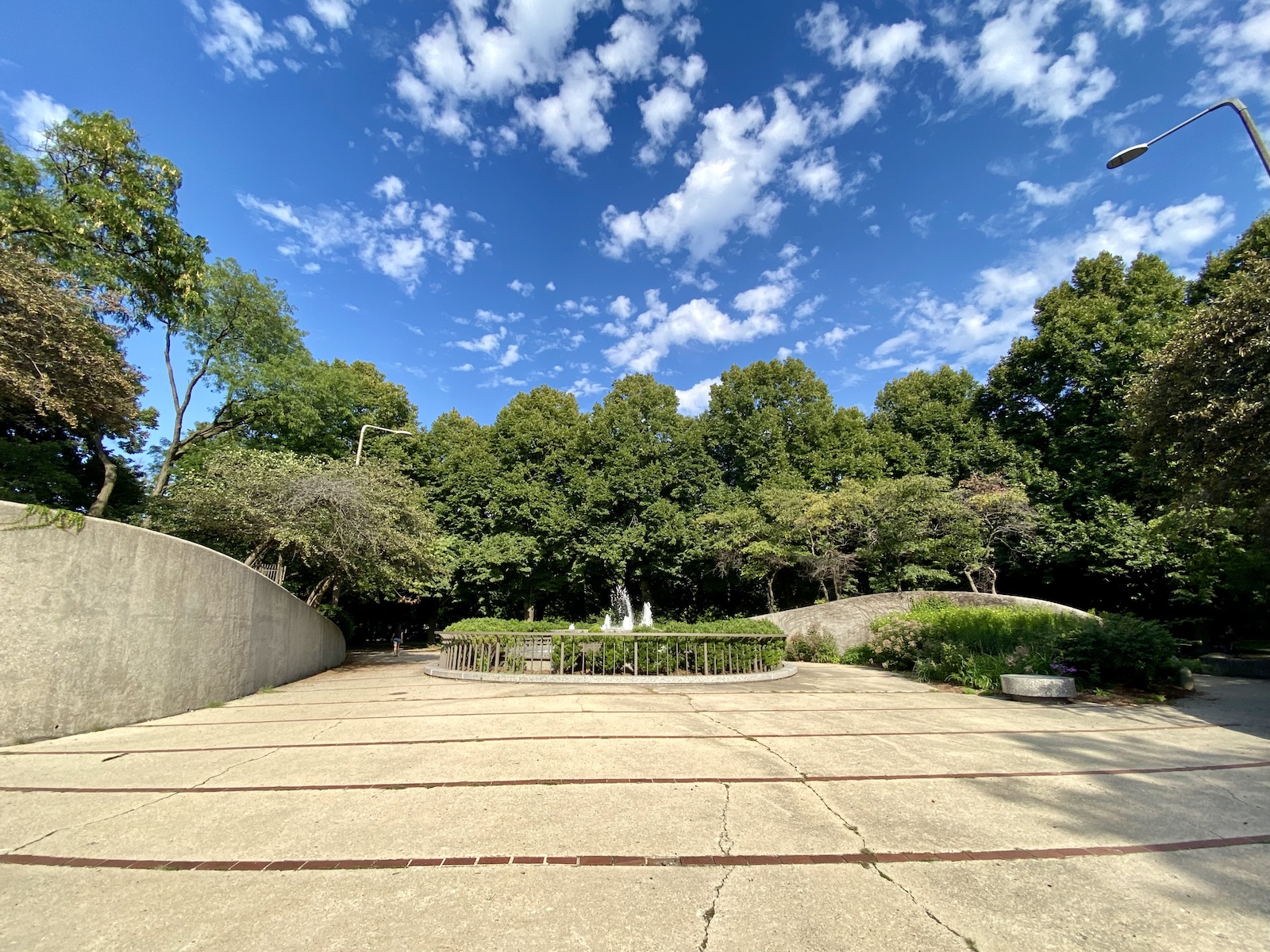 A fountain is centered surrounded by concrete curved walls and concrete paths with trees lined in the distance.